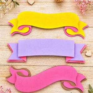 Puede incluir: Tres pancartas de papel en capas en amarillo, morado y rosa, con detalles en marrón y rosa. Las pancartas están dispuestas sobre una superficie de madera clara, con flores decorativas y corazones de madera esparcidos.