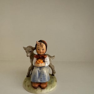 May include: A ceramic figurine of a young girl in a blue dress and red headscarf, holding a bundle of flowers. A gray goat stands behind her. The figurine is on a green and brown base.