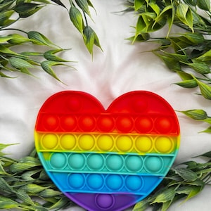 May include: A heart-shaped, rainbow-colored pop-it toy. The toy features a series of circular bubbles in red, orange, yellow, green, blue, and purple. The toy is surrounded by green foliage.