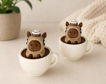 May include: Two ceramic coffee cups with a brown liquid inside, each holding a brown animal figurine with a white pastry on its head. The cups are white, and the background is neutral with a plant and a knitted blanket.