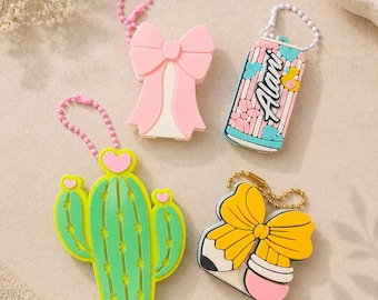 May include: Four colorful keychains: a pink bow, a can with floral design, a green cactus with yellow accents, and a pencil with a yellow bow. Each keychain has a chain for attachment.