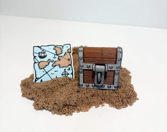 May include: A miniature treasure chest and a map on a bed of sand. The chest is brown with silver accents and a latch. The map is blue with brown landmasses and a ship illustration, evoking a pirate theme.
