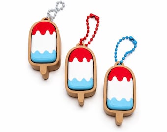 May include: Three popsicle-shaped keychains featuring a tan frame and a red, white, and light blue layered design. Each keychain includes a small stick at the bottom and a ball chain attachment in silver, red, or blue for hanging purposes.
