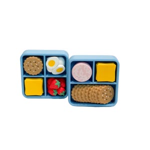 May include: Two light blue, divided containers filled with miniature food items. One container holds a round cookie, two egg halves, a yellow square, and strawberries. The other holds a slice of meat, a yellow square, and crackers.