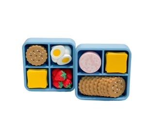 May include: Two light blue, divided containers filled with miniature food items. One container holds a round cookie, two egg halves, a yellow square, and strawberries. The other holds a slice of meat, a yellow square, and crackers.
