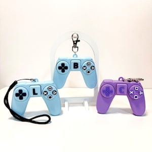 May include: Three game controller keyrings in pastel colours. Two are light blue, one with the letter "B" and the other with the letter "L". The third is purple with the letter "C". Each has a black strap or silver keyring.