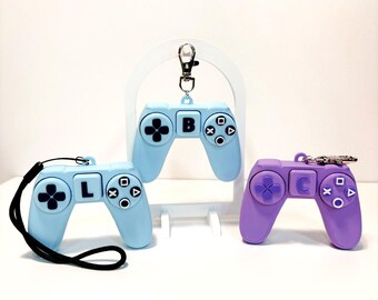 May include: Three colorful game controller keychains. Two are light blue, one with the letter "B" and the other with the letter "L". The third is purple with the letter "C". Each has a black strap or silver keychain.