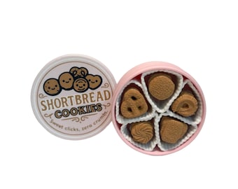 May include: A pink round box of shortbread cookies with a white interior, containing five different shapes of cookies. The lid is off, revealing the cookies. The lid has the text "Shortbread Cookies" and "Sweet clicks, zero crumbs".