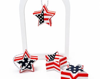 May include: Four star-shaped ornaments featuring a red, white, and blue pattern with white stars on a blue field and red and white stripes. Each star has a layered, three-dimensional design and includes a silver-toned ball chain for hanging.