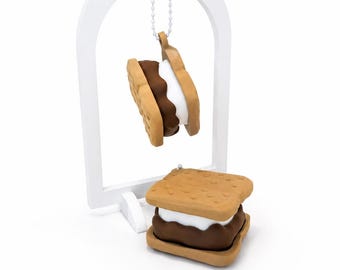 May include: A white arched display stand holds a s'more-shaped charm suspended by a silver ball chain. A second s'more-shaped object rests at the base of the stand. Both items feature tan graham cracker layers, a brown chocolate center, and white marshmallow.