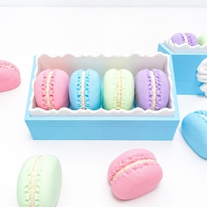 May include: A light blue rectangular container with a white scalloped edge holds four pastel-colored macaron-shaped objects in pink, blue, green, and purple. Additional individual macaron-shaped objects in pink, green, and blue are scattered around the container.