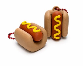 May include: Two hot dog keychains featuring a tan bun, a brown sausage, and a bright yellow wavy line of mustard. Each hot dog is attached to a red ball chain, with one positioned horizontally and the other standing vertically on its end.