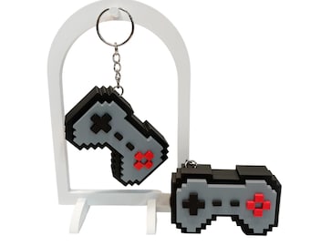 May include: Two pixelated video game controller keychains. One hangs from a silver keyring, the other rests below. Both are black and gray with red accents. They are displayed on a white stand.