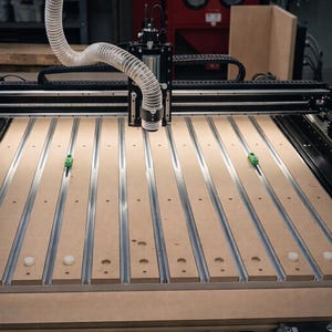 Puede incluir: Una máquina fresadora CNC con marco de metal negro, una base de MDF marrón claro con ranuras en T de aluminio paralelas y un cabezal central equipado con una manguera de aspiración blanca flexible y dos abrazaderas de color verde.