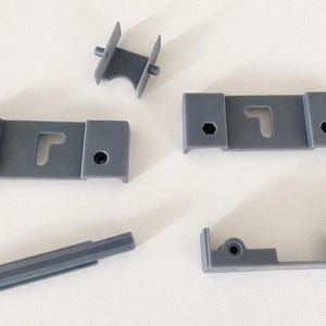 Pode incluir: Uma variedade de componentes de hardware de móveis de plástico cinza. As peças incluem suportes retangulares com recortes em forma de fechadura, um pequeno conector e um trilho deslizante. O hardware é projetado para montagem ou reparo de móveis.