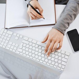 Op de afbeelding: Bovenaanzicht van een witte tafel met een wit toetsenbord, een smartphone en een open notitieboek. De handen van een persoon zijn zichtbaar, de ene typt op het toetsenbord en de andere schrijft in het notitieboek met een pen. De smartphone heeft een zwart scherm.