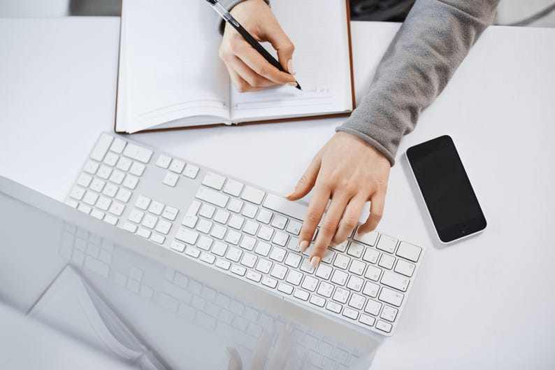 Op de afbeelding: Bovenaanzicht van een witte tafel met een wit toetsenbord, een smartphone en een open notitieboek. De handen van een persoon zijn zichtbaar, de ene typt op het toetsenbord en de andere schrijft in het notitieboek met een pen. De smartphone heeft een zwart scherm.