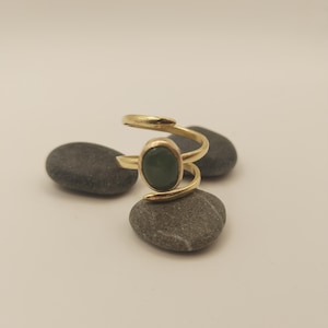Può includere: Un anello dorato con una pietra ovale verde scuro. La fascia dell'anello si avvolge attorno al dito, creando un design aperto unico. L'anello è esposto su una pietra grigia, evidenziandone l'elegante semplicità.