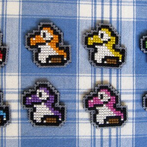 May include: Eight pixel art dinosaur figures in red, orange, yellow, green, blue, purple, pink, and rainbow colours. Each has a white belly, brown feet, and a grey outline. They are arranged on a blue and white checkered surface, reminiscent of a tablecloth.