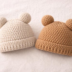 Może przedstawiać: Dwie ręcznie robione czapki dla niemowląt na szydełku. Jedna jest w kolorze jasnobeżowym, a druga w ciepłym brązie. Każda czapka ma dwa małe, okrągłe akcenty w kształcie uszu na górze. Czapki są wykonane z teksturowanego ściegu.