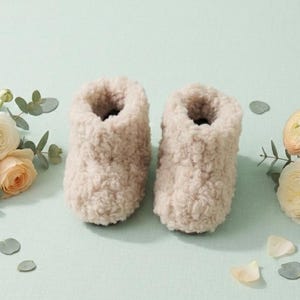 May include: A pair of beige, high-top booties crafted from a thick, textured, fleece-like material. The booties are positioned side-by-side, flanked by two small bouquets of pale peach and cream-colored ranunculus flowers with green eucalyptus leaves.