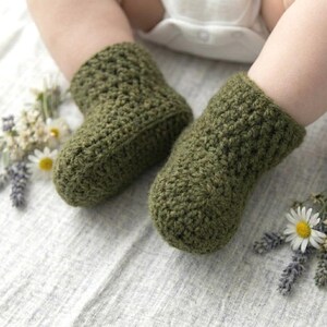 May include: A pair of olive green crochet booties with a textured stitch pattern. The booties are worn on feet and are surrounded by small sprigs of white daisies and purple lavender flowers on a light-colored, textured fabric surface.