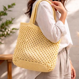 May include: A pale yellow crochet tote bag featuring an open diamond-shaped mesh pattern throughout the main body and a solid, tightly stitched base. The bag includes two integrated shoulder straps and is constructed from a soft yarn material.