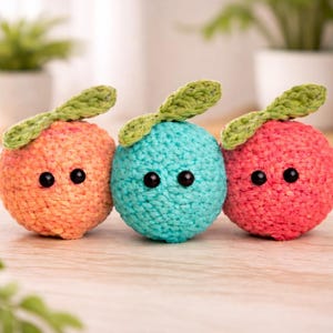 May include: Three handmade crochet fruit toys in orange, teal, and red. Each has a green leaf and black button eyes, giving them a whimsical appearance. The toys are arranged on a light wooden surface, suggesting a playful theme.