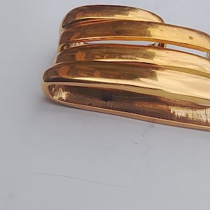 May include: A gold-toned metal scarf slide or brooch featuring three stacked, smooth, curved horizontal bars. The piece has a polished, reflective finish with a hollow interior structure and a solid, continuous loop design on the side.