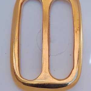 May include: A rectangular gold-toned metal belt buckle slider featuring rounded corners and a central vertical bar that divides the interior space into two equal openings. The surface has a smooth, polished finish with a reflective metallic sheen.