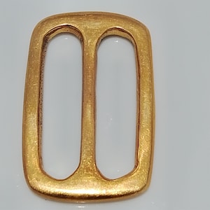May include: A gold-colored metal buckle with a rectangular shape and two oval openings. The buckle has a slightly textured surface and rounded edges. The design is simple and elegant, suitable for belts or other accessories.