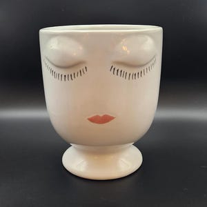 May include: A white ceramic planter shaped like a human head with closed eyes, long black eyelashes, and small pink lips. The vessel features a smooth, glossy finish and sits on a circular pedestal base, creating a sculptural container for plants.