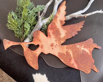 May include: A rustic metal angel silhouette with a trumpet, featuring a heart-shaped cutout. The angel has a weathered, orange-brown finish, suggesting an aged appearance. Green sprigs and a light-colored branch are placed behind the angel.