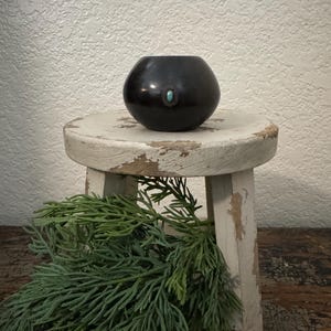 May include: A small, round, black ceramic bowl with a turquoise stone accent. The bowl sits on a distressed white stool, with green foliage in the foreground. The bowl is approximately 4 inches in diameter.