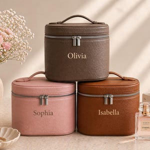 May include: Three personalised cosmetic cases in shades of brown and pink are stacked. Each case has a top handle, a silver zip, and a name: Olivia, Sophia, and Isabella. A vase of pink flowers and perfume are in the background.