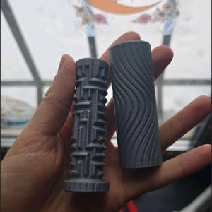 May include: Two gray cylindrical objects with textured surfaces. One has a geometric pattern, the other a wavy design. The objects are held in a hand, with a decorative sticker featuring a moon and mushrooms in the background.