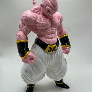 May include: A pink action figure of a muscular character with a distinctive head shape, white pants, and black boots. The figure has black armbands with gold trim and a black belt with a gold "M" buckle. The figure is from the Dragon Ball Z series.