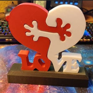 May include: A 3D-printed sculpture featuring a red and white heart puzzle with interlocking shapes. The word "LOVE" is displayed in red and white letters on a black base. The sculpture is a symbol of love and connection.