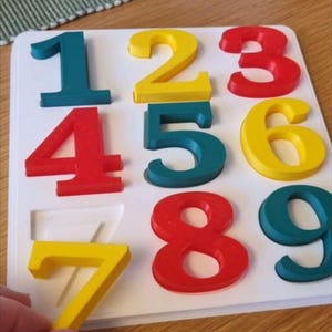 Könnte beinhalten: Ein farbenfrohes Holz-Zahlenpuzzle mit den Zahlen 1 bis 9. Die Zahlen sind in verschiedenen Farben gehalten: teal, gelb und rot. Das Puzzle befindet sich auf einer weißen Basis. Eine Hand hält die gelbe Zahl 7.