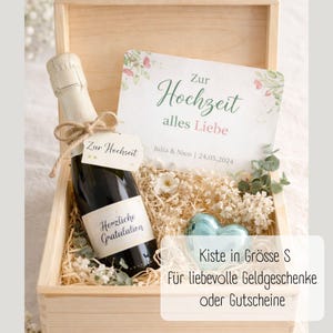 May include: A wooden gift box with a bottle of champagne, a greeting card, and decorative elements. The card reads "Zur Hochzeit alles Liebe" and includes a date. The champagne bottle has a tag that says "Zur Hochzeit" and "Herzliche Gratulation".