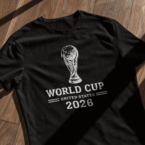 May include: Black t-shirt with a white graphic of the World Cup trophy. The text "WORLD CUP UNITED STATES 2026" is printed below the trophy in a bold, sans-serif font. The shirt is displayed on a wooden surface.