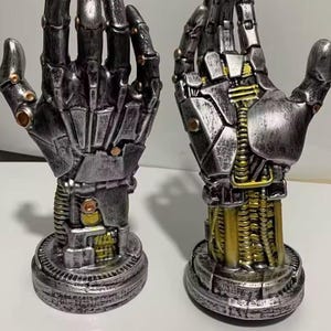 Könnte beinhalten: Zwei metallische Roboterhand-Skulpturen. Die Hände sind silber und schwarz mit goldenen Akzenten und weisen komplizierte mechanische Details auf. Jede Hand ist auf einem kreisförmigen Sockel montiert.