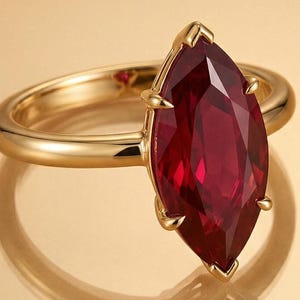 May include: A gold ring featuring a large, marquise-cut ruby gemstone. The ruby is a deep red color and is set in a gold band with a polished finish. The ring is displayed on a reflective surface.