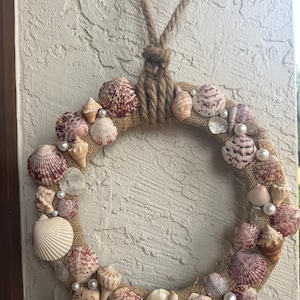 May include: A circular wreath made of burlap, adorned with various seashells in shades of white, pink, and brown. Faux pearls are interspersed among the shells. A rope loop is attached for hanging.