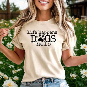 K&ouml;nnte beinhalten: Cremefarbenes T-Shirt mit dem schwarzen Text "life happens DOGS help". Das Wort "DOGS" hat ein Pfotenabdruck-Design. Ein l&auml;ssiges Kleidungsst&uuml;ck f&uuml;r den Alltag.