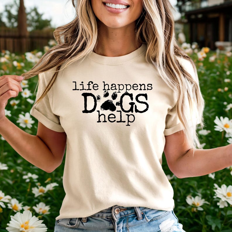 K&ouml;nnte beinhalten: Cremefarbenes T-Shirt mit dem schwarzen Text "life happens DOGS help". Das Wort "DOGS" hat ein Pfotenabdruck-Design. Ein l&auml;ssiges Kleidungsst&uuml;ck f&uuml;r den Alltag.