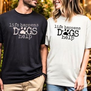 K&ouml;nnte beinhalten: Zwei kurz&auml;rmelige T-Shirts, eines in Schwarz und eines in Off-White, mit dem Text "life happens DOGS help" auf der Vorderseite. Der Text ist in einer Used-Schriftart mit einem Pfotenabdruck-Motiv. Die Shirts werden von zwei Personen getragen.