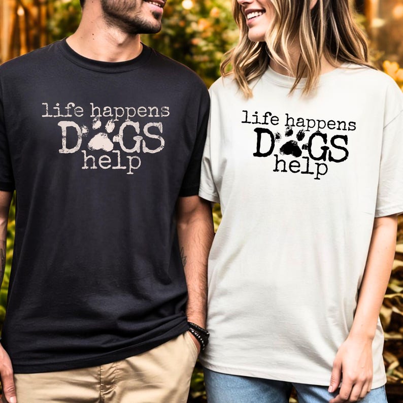K&ouml;nnte beinhalten: Zwei kurz&auml;rmelige T-Shirts, eines in Schwarz und eines in Off-White, mit dem Text "life happens DOGS help" auf der Vorderseite. Der Text ist in einer Used-Schriftart mit einem Pfotenabdruck-Motiv. Die Shirts werden von zwei Personen getragen.