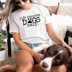 K&ouml;nnte beinhalten: Beiges T-Shirt mit schwarzem Aufdruck "life happens DOGS help". Das Wort "DOGS" enth&auml;lt einen Pfotenabdruck. Ein braun-wei&szlig;er Hund ist im Vordergrund.