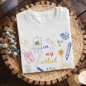 Könnte beinhalten: Weißes T-Shirt mit farbenfrohen Illustrationen zum Thema Künstler und dem Text "in my artist era". Das Design umfasst Pinsel, Farbtuben, einen Bleistift und eine Staffelei mit Leinwand. Das T-Shirt wird auf einem geflochtenen Korb präsentiert.